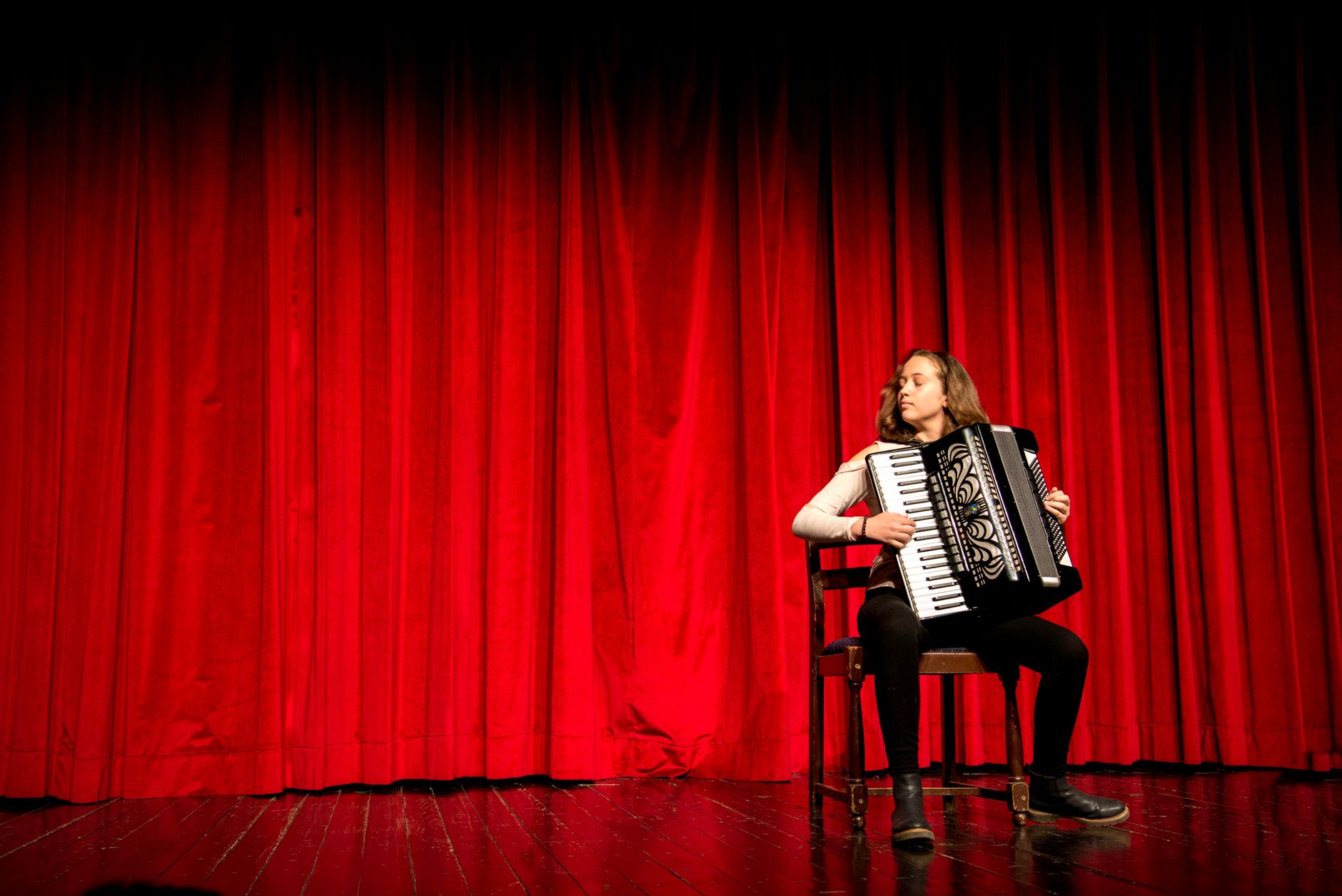Une musicienne joue de l’accordéon, assise sur une chaise devant un grand rideau rouge sur une scène de théâtre.